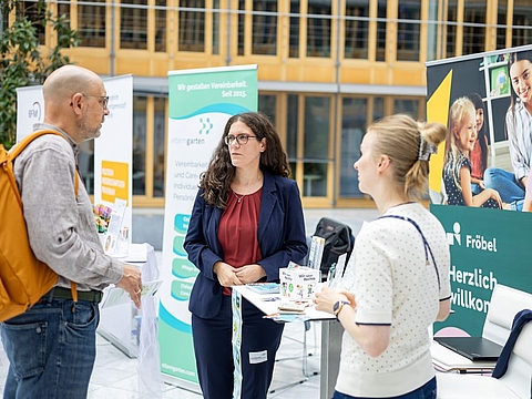 Drei erwachsene Personen unterhalten sich an einem Fröbel-Informationsstand. Eine Frau mit dunklen Haaren steht zwischen einem Mann mit orangem Rucksack und einer Frau mit hellem Oberteil. Auf dem Tisch liegen Broschüren und weitere Informationsmaterialien. Im Hintergrund sind Roll-up-Banner mit Fröbel-Motiven sowie Bildern von Kindern und Erwachsenen zu sehen.