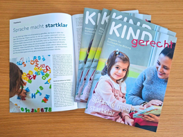 Mehrere Ausgaben des Magazins „KINDgerecht“ mit dem Titel „Sprache macht startklar“ vom September 2026 liegen auf einem Holztisch; eine Ausgabe ist aufgeschlagen.