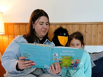 Eine junge Frau mit langen, dunklen Haaren und einer hellblauen Jeansjacke sitzt auf einem Sofa vor einer holzvertäfelten Wand. Sie hält ein türkischsprachiges Bilderbuch mit türkisfarbenem Einband aufgeschlagen. Der Titel lautet „Küçük Biz Anaokulunda“. Neben ihr sitzt ein Mädchen in einem türkisfarbenen Pullover, das aufmerksam in das Buch schaut. Hinter den beiden steht ein schwarzes Plüschtier mit gelbem Schnabel. Links im Bild ist eine Lampe zu sehen.