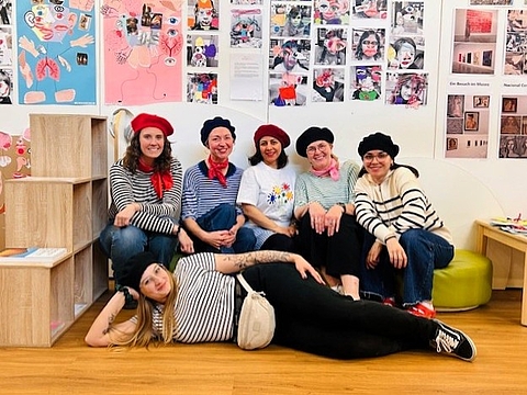 Gruppe von Erwachsenen in einem Ausstellungsraum mit Kunstwerken an den Wänden, einige sitzen, andere liegen entspannt auf dem Boden.