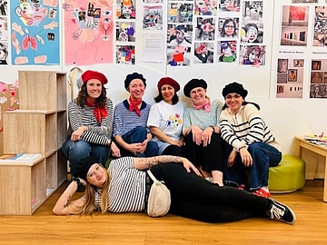 Gruppe von Erwachsenen in einem Ausstellungsraum mit Kunstwerken an den Wänden, einige sitzen, andere liegen entspannt auf dem Boden.