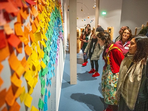 Teilnehmende stehen vor einer Wand voller Klebezettel in Regenbogenfarben, auf denen Kinderwünsche für die Zukunft stehen.
