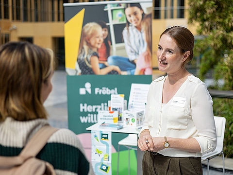 Eine Frau in einer weißen Bluse steht an einem Fröbel-Informationsstand und spricht mit zwei weiteren Personen. Im Hintergrund sind Banner, Informationsmaterialien und Bilder von Kindern und Erwachsenen zu sehen.