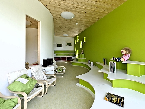  Ein heller und freundlicher Innenraum eines Kindergartens mit einer wellenförmigen, stufenartigen Sitz- und Spiellandschaft in Weiß und Hellgrün. An der Wand sitzt eine Handpuppe. Der Raum ist minimalistisch eingerichtet mit Holzboden und einer grünen Wand.