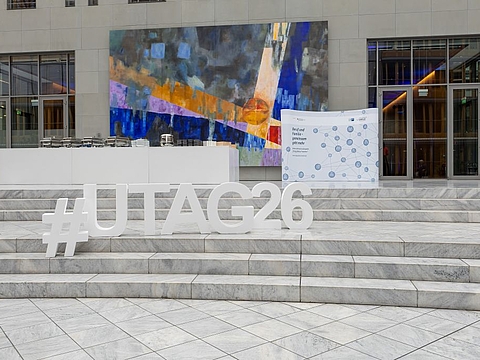 Auf einer breiten Marmortreppe stehen große weiße Buchstaben mit der Aufschrift „#UTag26“. Dahinter befinden sich ein Buffet, ein farbenfrohes Wandbild und ein Aufsteller des Netzwerks „Erfolgsfaktor Familie“.