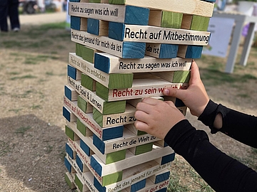 Kinderhände ziehen einen Holzklotz aus einem großen Kinderrechte-Jenga-Turm im Freien. Auf den Bausteinen stehen verschiedene Kinderrechte, darunter „Recht auf Mitbestimmung“ und „Recht zu sagen, was ich will“.