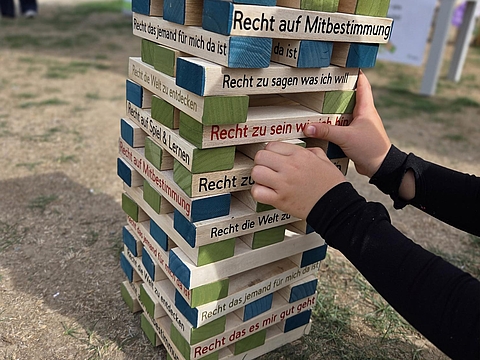 Kinderhände ziehen einen Holzklotz aus einem großen Kinderrechte-Jenga-Turm im Freien. Auf den Bausteinen stehen verschiedene Kinderrechte, darunter „Recht auf Mitbestimmung“ und „Recht zu sagen, was ich will“.