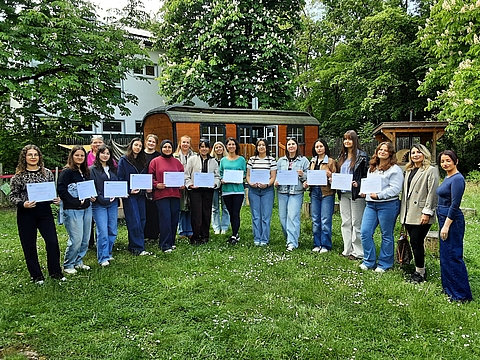 Gruppenfoto im Freien auf einer grünen Wiese mit weißen Blüten. Rund 18 Personen stehen nebeneinander und halten überwiegend helle Blätter oder Dokumente vor sich. Die Frauen tragen unterschiedliche Kleidung, darunter Jeans, Pullover und Jacken. Im Hintergrund sind ein weißes Gebäude, große blühende Bäume und eine hölzerne Hütte mit rundem Dach zu sehen.