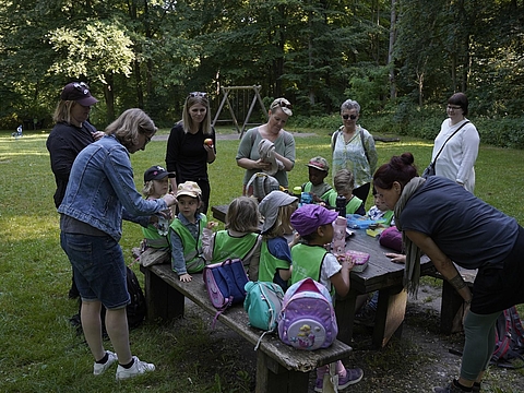 Kinder und pädagogische Fachkräfte sitzen an einem Rastplatz im Wald und frühstücken gemeinsam.