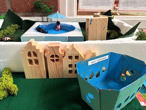 Modell einer Stadtlandschaft aus Holz, Karton und Naturmaterialien mit Häusern, Grünflächen und einem kleinen Schwimmbecken.