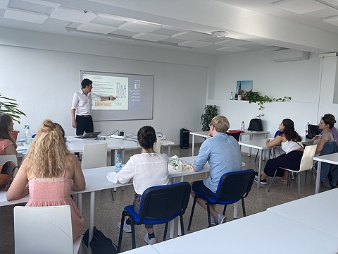 Ein Mann steht vorne im Seminarraum und zeigt auf eine Präsentation, während mehrere Personen des Teams Personalgewinnung an weißen Tischen sitzen und zuhören.
