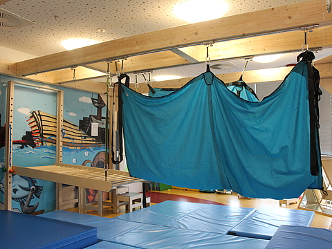 Foto vom Innenraum des FRÖBEL-Kindergartens Hamburger Meile in Hamburg. Ein Bewegungsraum mit großen Dachbalken, an denen Kletterelemente und ein großes Schwungtuch hängen