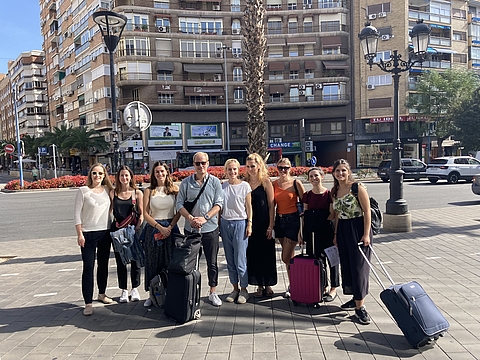 Neun Mitglieder des Teams Personalgewinnung von Fröbel stehen mit Koffern in sommerlicher Kleidung auf einem Platz in Alicante, Spanien. Im Hintergrund mehrstöckige Wohnhäuser, Palmen und Straßenszene bei Tageslicht.