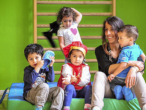 [Translate to Englisch:] Eine Erzieherin und mehrere Kinder sitzen im Bewegungsraum auf Sportmatten. Im Hintergrund ist eine Sprossenwand.