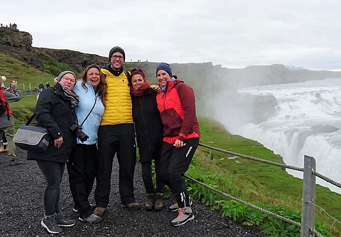 [Translate to Englisch:] Foto von einer Gruppe pädagogischer Fachkräfte auf Erasmus+ Bildungsreise in Island vor einem Wasserfall. Sie lachen und winken in die Kamera.