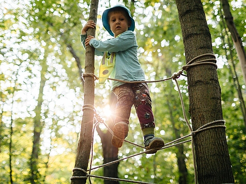 Kind im Wald auf einem Seil, das zwischen Baumstämme gespannt ist. (FRÖBEL forstet auf: Eine Initiative für mehr Nachhaltigkeit)