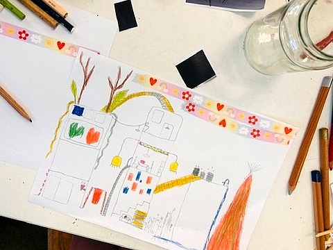 Kinderzeichnung einer Stadt mit Häusern, Wegen, Pflanzen und farbigen Elementen auf einem Tisch mit Stiften.