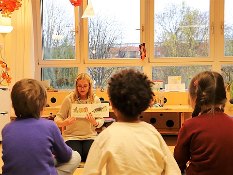 Sprachförderkraft Natalie sitzt mit Kindern im Kreis. Sie hält das Buch hoch und erzählt. Die Kinder lauschen aufmerksam.