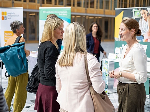 Eine Frau in weißer Bluse spricht mit zwei weiteren Frauen in einem Veranstaltungsfoyer. Im Hintergrund stehen Informationsstände mit Roll-up-Bannern, Broschüren und Bildern von Kindern und Erwachsenen.
