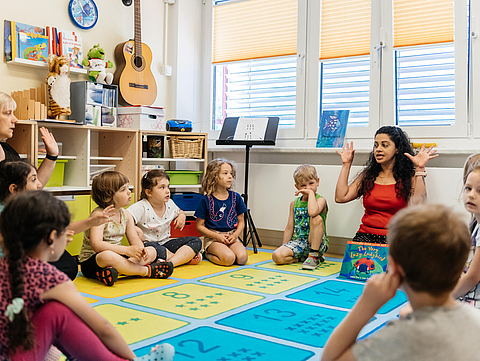 Foto von zwei Erzieherinen, die mit Kindern im Kindergarten in einem Kreis sitzen. Sie sprechen und machen passende Gesten dazu. Die Kinder schauen aufmerksam zu.