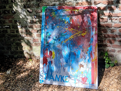 Ein großes Gemälde lehnt im Freien an einer alten Backsteinwand auf Holzhäckseln. Das Bild ist abstrakt gestaltet, mit kräftigen Flächen in Blau, Rot, Gelb und Grün, die sich mit weißen Farbakzenten und Spritzern mischen. Am unteren Rand steht ein blauer Schriftzug, der teilweise verdeckt ist. Im Hintergrund wächst Efeu an der Mauer, und Schatten von Bäumen fallen auf die Wand.