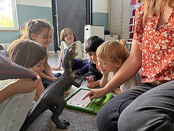 Fünf Kinder sitzen im Kindergarten gemeinsam mit einer pädagogischen Fachkraft auf dem Boden um ein Tablet und einen großen Spielzeug-Dinosaurier.