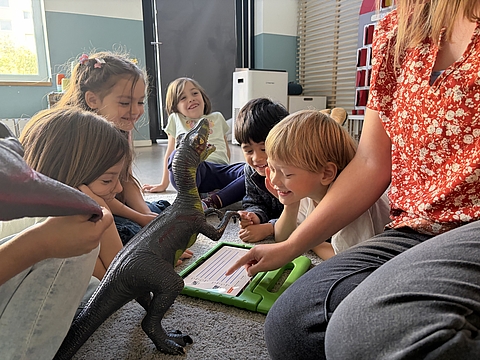 Fünf Kinder sitzen im Kindergarten gemeinsam mit einer pädagogischen Fachkraft auf dem Boden um ein Tablet und einen großen Spielzeug-Dinosaurier.