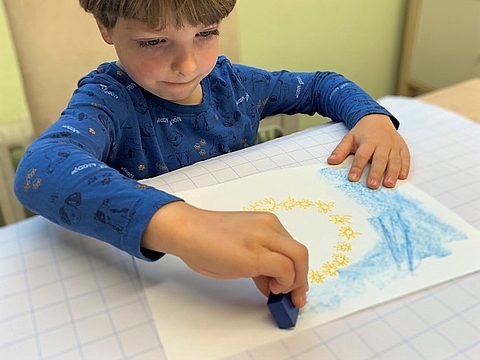 Kind sitzt am Tisch und malt eine Europaflagge auf Papier