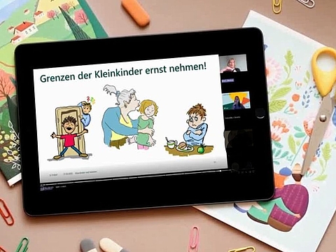 Das Bild zeigt ein schwarzes Tablet, das auf einem Schreibtisch liegt – umgeben von buntem Büromaterial (Papier, Schere, Stifte, Papierklammern) und einem kindlich gestalteten Block mit Blumenmuster.  Auf dem Tablet läuft eine Online-Präsentation der Fröbel-Elternakademie. Auf dem Bildschirm ist eine Folie mit dem Titel „Grenzen der Kleinkinder ernst nehmen!“ zu sehen. Die Folie zeigt drei illustrierte Szenen:  links: ein Kind, das wütend die Tür zuschlägt  in der Mitte: ein Kind, das sich von einer Erwachsenen distanzieren möchte, aber von ihr festgehalten wird  rechts: ein trotzig blickendes Kind am Esstisch, das offenbar nicht essen möchte  Rechts auf dem Bildschirm sind kleine Kacheln mit der Referentin Marion Koch, Moderatorin Christin Schröder und weiteren Teilnehmenden der Online-Veranstaltung zu sehen.