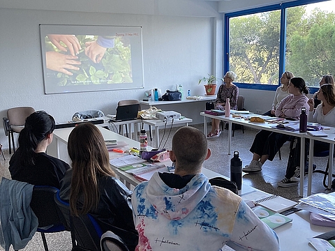 Mehrere junge Erwachsene sitzen in einem hellen Seminarraum und schauen gemeinsam ein Video, das an die Wand projiziert wird. Im Bild sind Hände im Gemüsebeet zu sehen. Eine Fröbel-Mitarbeitende sitzt vorne rechts.