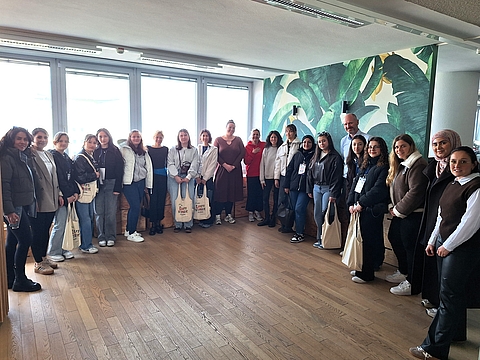 Gruppenfoto in einem hellen Raum mit Holzboden und großen Fenstern. Rund 20 Personen stehen vor einer Wand mit großflächigem grünem Blattmuster. Die jungen Frauen tragen Taschen aus Stoff, auf denen „Laut stark“ steht. Die Gruppe steht dicht beieinander und blickt in die Kamera.