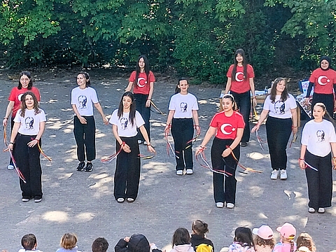 Rund zwölf junge Frauen tanzen im Freien auf einem Platz mit Schotter, vor einer grünen Hecke. Sie tragen rote T-Shirts mit einem weißen Halbmond und Stern oder weiße T-Shirts mit dem Porträt eines Mannes und schwarzen Hosen. Jede Tänzerin hält bunte Bänder in den Händen. Im Vordergrund sitzen kleine Kinder auf dem Boden und schauen zu.