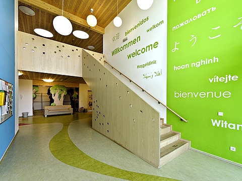 Ein heller Eingangsbereich eines Kindergartens mit einer Holztreppe und einer Wand, auf der das Wort „Willkommen“ in verschiedenen Sprachen steht. Der Boden ist mit einem geschwungenen grünen Muster versehen.