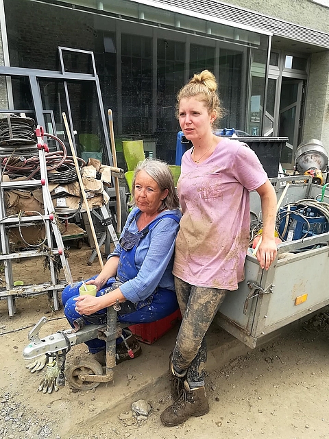 Zwei Frauen in verschlammten Arbeitssachen und Gummistiefeln sitzen bzw. stehen an einem Anhänger vor einem zerstörten Haus und machen eine kleine Pause