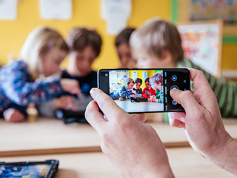 Foto von Kinder in einem Kindergarten, die an einem Tisch sitzen und ein Tablet benutzen. Im Vordergrund halten zwei Hände einer erwachsenen Person ein Smartphone in der Hand und fotografiert die Szene 