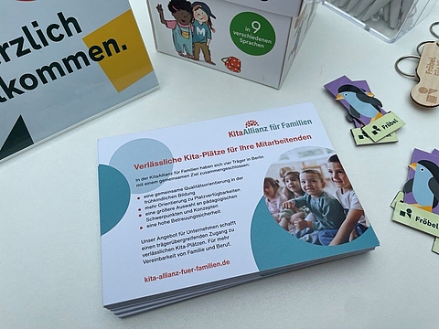 Auf einem Tisch liegen eine Broschüre der KitaAllianz für Familien mit der Überschrift „Verlässliche Kita-Plätze für Ihre Mitarbeitenden“, ein Aufsteller mit dem Hinweis „In neun verschiedenen Sprachen“ sowie mehrere Fröbel-Schlüsselanhänger und weitere Informationsmaterialien. Die Broschüre zeigt ein Foto von Kindern.