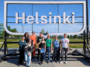 Eine Gruppe von zehn Erwachsenen steht lächelnd vor einem großen, weißen, dreidimensionalen Schriftzug, der „Helsinki“ buchstabiert. Der Himmel ist blau und klar. Die Personen stehen verstreut vor und zwischen den Buchstaben.