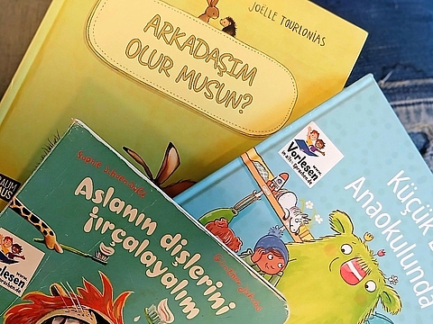 Drei türkischsprachige Bilderbücher liegen übereinander auf einer dunklen Unterlage. Ganz oben liegt ein gelbes Buch mit dem Titel „Arkadaşım olur musun?“ von Joëlle Tourlonias, auf dem zwei Tiere abgebildet sind. Darunter liegt ein grünes Buch mit dem Titel „Aslanın dişlerini fırçalayalım“ von Sophie Schoenwald, auf dem ein Löwe mit roter Mähne zu sehen ist. Rechts liegt ein hellblaues Buch mit dem Titel „Küçük Biz Anaokulunda“, auf dem ein grünes Monster und Kinder abgebildet sind. Auf den Büchern kleben Aufkleber des Projekts „Vorlesen in allen Sprachen“.