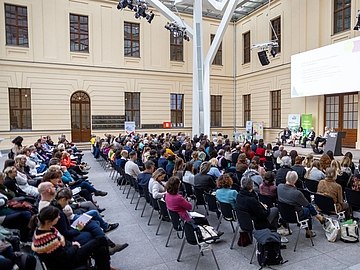Foto vom Glashof des Jüdischen Museums mit vielen Menschen, die in Stuhlreihen sitzen und einer Diskussion auf der Bühe zuhören.  
