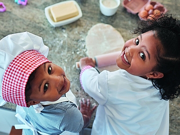 Zwei Kinder in weißen Kochschürzen und Kochmützen backen gemeinsam. Das jüngere Kind trägt eine rot-weiß karierte Mütze und schaut in die Kamera, das ältere Kind hält ein Nudelholz und lächelt. Auf dem Tisch liegen Teig, Butter, Eier und bunte Ausstechformen.