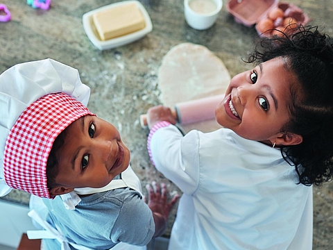 Zwei Kinder in weißen Kochschürzen und Kochmützen backen gemeinsam. Das jüngere Kind trägt eine rot-weiß karierte Mütze und schaut in die Kamera, das ältere Kind hält ein Nudelholz und lächelt. Auf dem Tisch liegen Teig, Butter, Eier und bunte Ausstechformen.