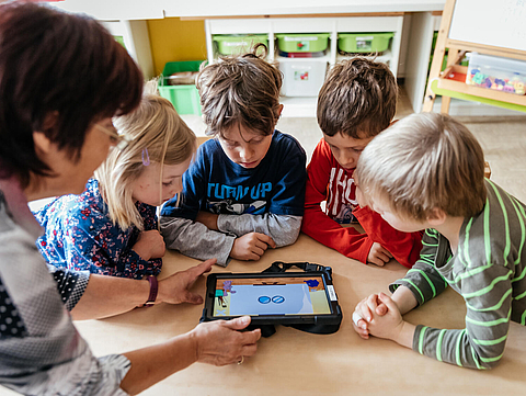 Foto von einer Erzieherin und vier Kindern an einem Tisch. Sie benutzen gemeinsam eine App auf einem Tablet.