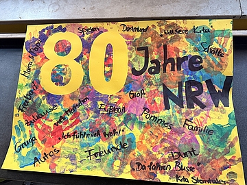  Buntes Kinderkunstwerk zum 80. Geburtstag von NRW mit großer „80“, dem Schriftzug „Jahre NRW“ und Begriffen wie Familie, Freunde, Fußball, Pommes und „Ich fühle mich wohl“. Entstanden im Fröbel-Kindergarten Sterntaler.