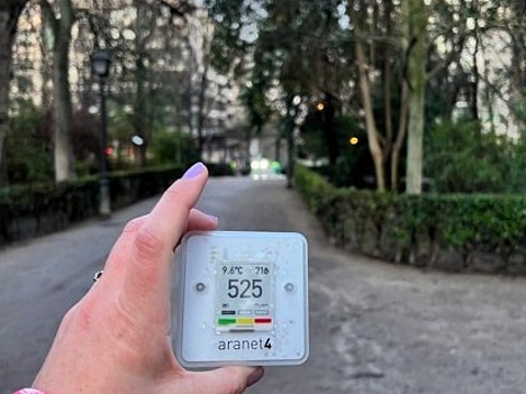 Hand hält ein digitales CO₂-Messgerät in einer begrünten Parkanlage mit Weg und Bäumen im Hintergrund.