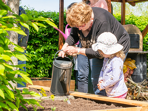  Eine Erwachsene und ein Kind gießen Pflanzen in einem Hochbeet im Garten. Die Szene spielt unter freiem Himmel, umgeben von grünen Blättern und Sträuchern.