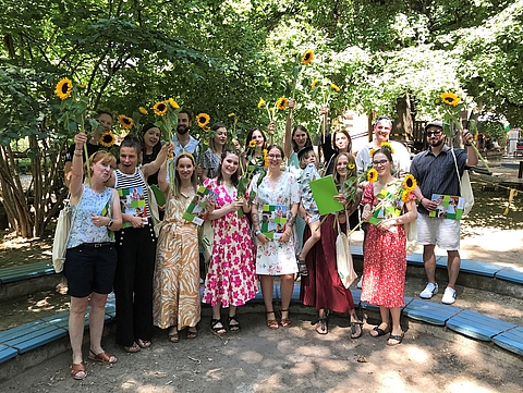 Gruppenfoto der Studierenden. Sie halten die Zeugnismappen in den Händen und halten große Sonnenblumen in die Höhe.