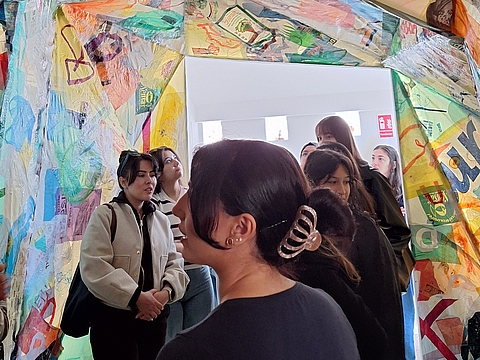 Blick durch einen Durchgang, dessen Rahmen mit bunten, collageartigen Materialien aus Folie und Papier beklebt ist. Im Vordergrund steht eine Frau mit dunklem, hochgestecktem Haar und einer Haarklammer, mit dem Rücken zur Kamera. Links und dahinter stehen mehrere junge Frauen, die aufmerksam zuhören und in den Raum schauen. Durch den Durchgang ist ein heller Raum mit Fenstern zu sehen.