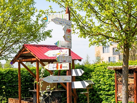Ein hölzerner Wegweiser mit mehreren Pfeilen, die verschiedene Orte und Entfernungen (z.B. Portugal, Spanien, Italien) anzeigen, steht inmitten eines grünen Gartens mit Bäumen und Sträuchern. Im Hintergrund ist ein Gebäude zu sehen.