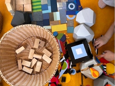 Verschiedene Bau- und Gestaltungsmaterialien wie Holzklötze, geometrische Formen, Stoffe und Kartonelemente auf dem Boden.