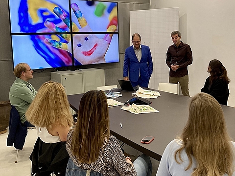 Mehrere Mitglieder des Teams Personalgewinnung sitzen um einen Konferenztisch. Zwei Männer – einer im blauen Anzug, einer im dunkelroten Hemd – stehen und sprechen vor der Gruppe. Im Hintergrund ein Monitor mit einem bunten Kinderhandabdruck mit aufgemaltem Gesicht.